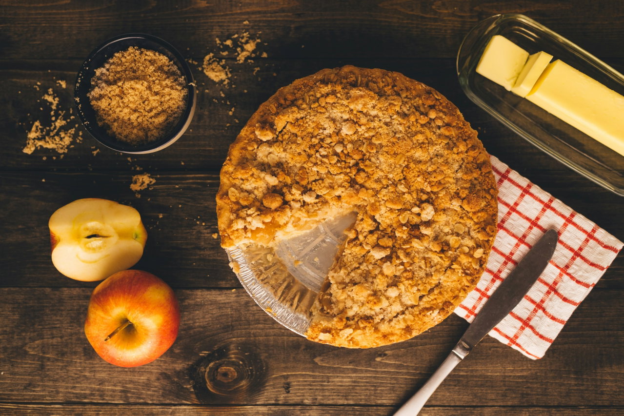 Apfel-Streuselkuchen auf einem Holztisch, daneben Äpfel, Butter und eine Schale braunen Zucker