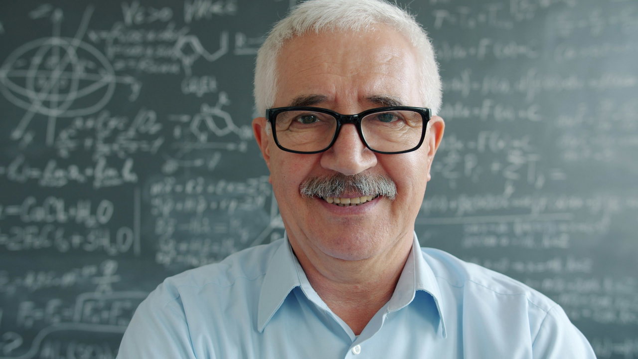 Älterer Mann mit weißem Haar, Schnurrbart und Brille lächelt vor einer Tafel mit Formeln