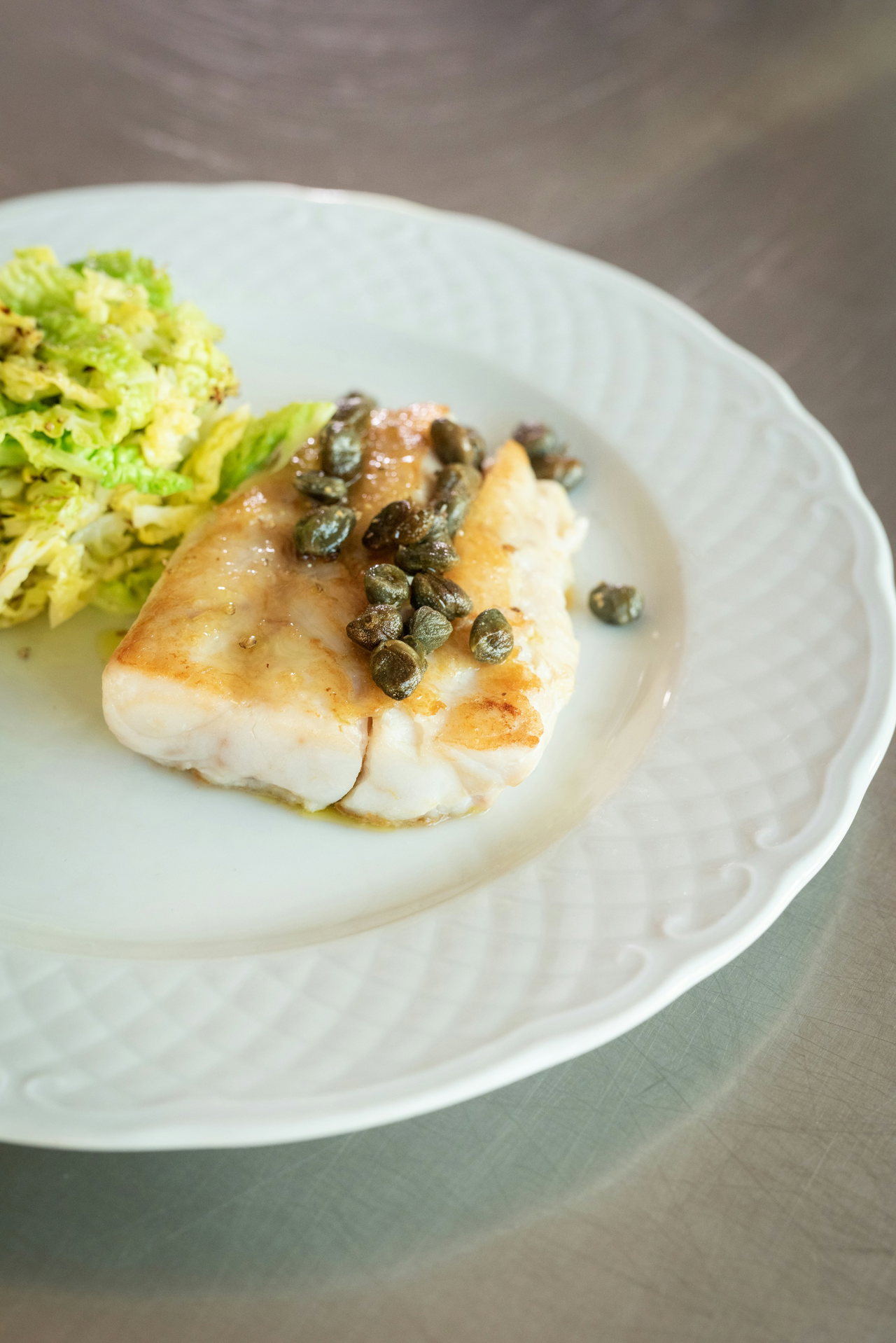 Gebratenes Fischfilet mit Kapern in Buttersauce auf weißem Porzellanteller, daneben Wirsinggemüse