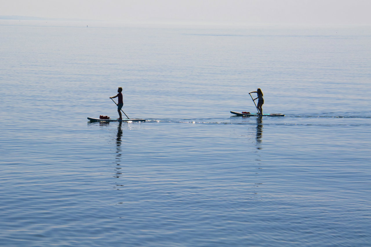 Zwei Stand-up-Paddler gleiten als Silhouetten über das spiegelglatte, hellblaue Meer an einem ruhigen Tag