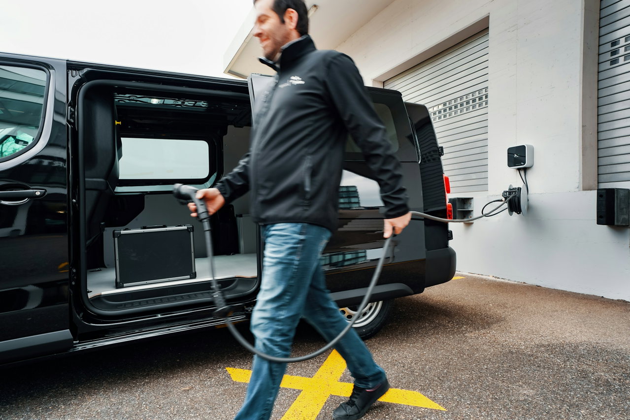 Handwerker zieht ein Ladekabel zu seinem dunklen Elektro-Transporter, der vor einer Werkhalle an der Wallbox lädt