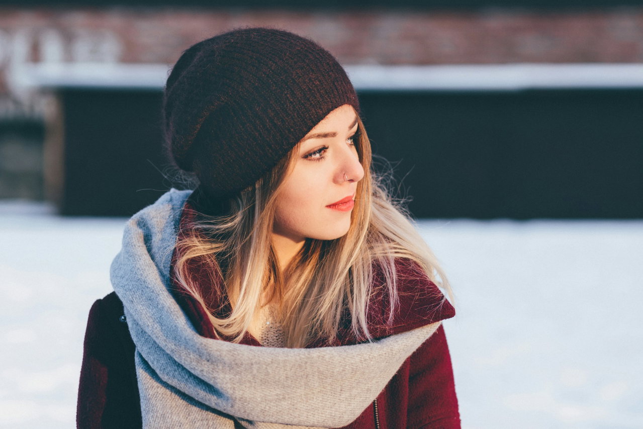 Frau mit schwarzer Strickmütze und grauem Schal über weinrotem Mantel im Schnee, Blick zur Seite