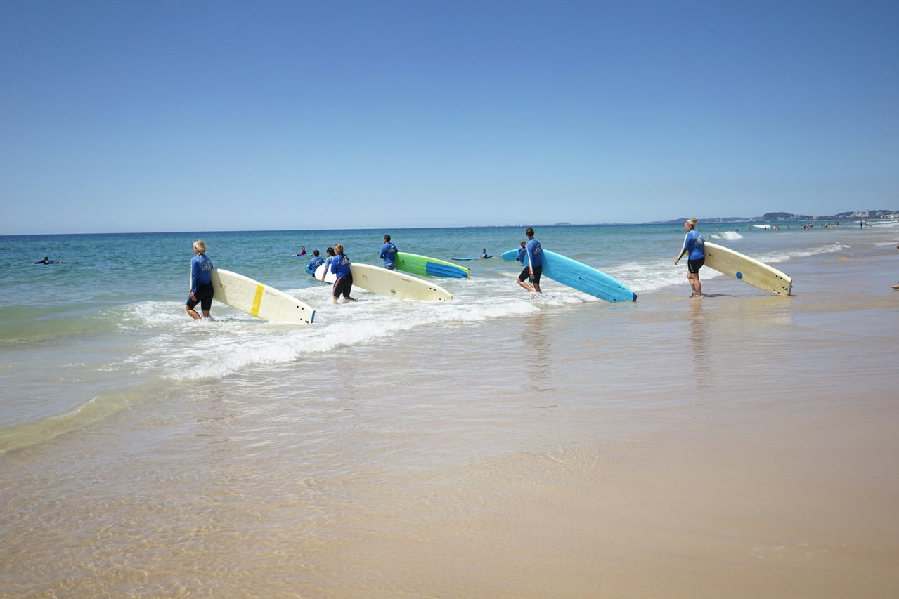 Surfschüler tragen ihre Bretter ins flache Wasser eines hellen Sandstrands unter blauem Himmel