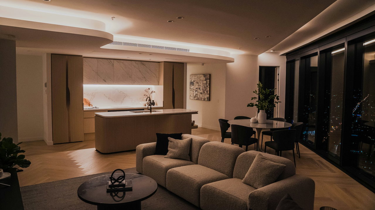 Großer Wohnraum am Abend mit Sofa, Essplatz und Kücheninsel unter indirekt beleuchteter Decke, Fischgrätparkett und raumhohe Fenster mit den Lichtern der Stadt.
