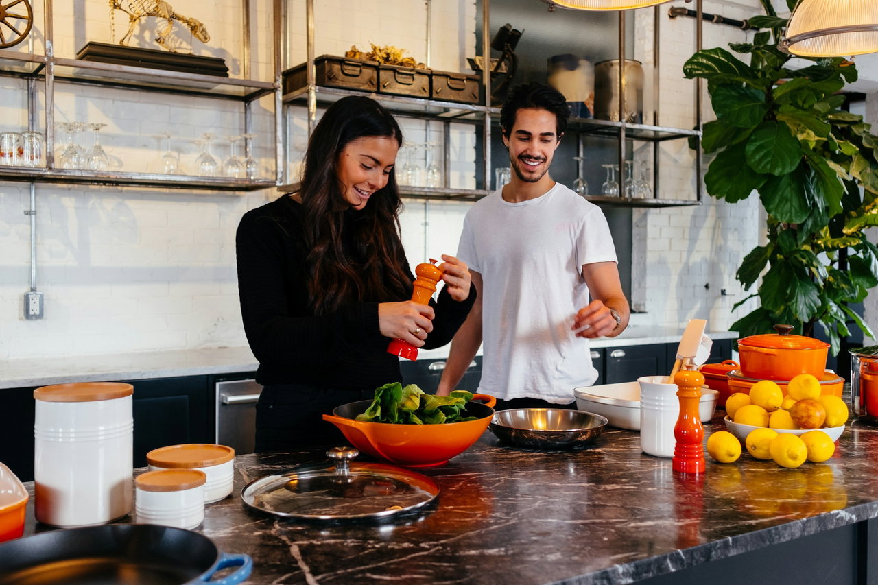Eine junge Frau und ein junger Mann kochen lachend an einer Kücheninsel mit dunkler Marmorplatte, Zitronen, orangefarbenen Töpfen und einer Salatschüssel, dahinter offene Regale und eine große Pflanze.
