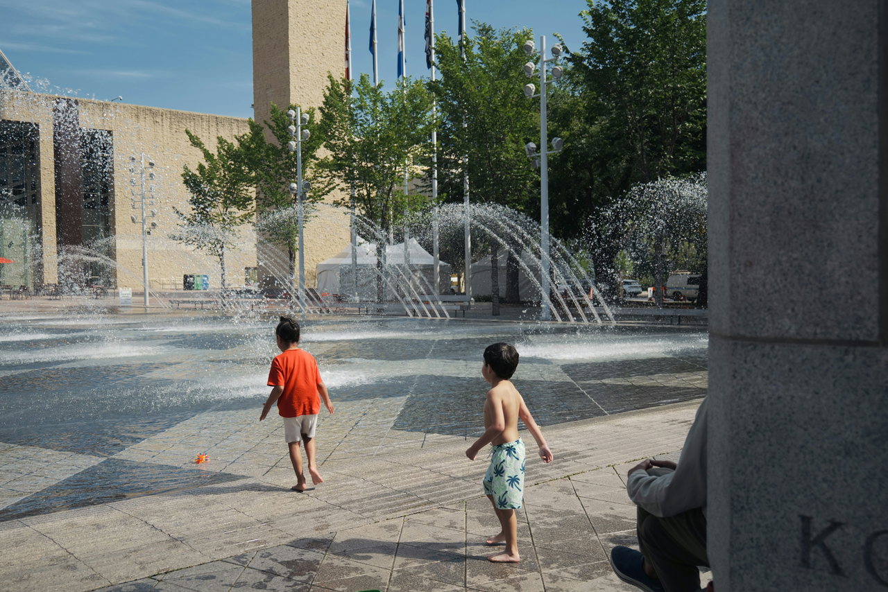 Zwei Kinder spielen an einem Wasserspiel auf einem sonnigen Stadtplatz zwischen aufsteigenden Fontänen