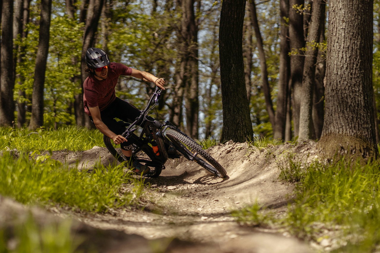 Mountainbiker mit Helm und weinrotem Shirt legt sich auf einem Waldtrail tief in eine Steilkurve