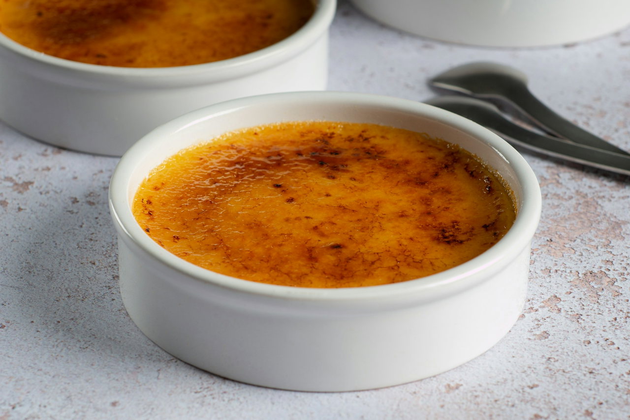 Crème brûlée mit karamellisierter Zuckerkruste in weißen Förmchen auf hellem Steintisch, daneben Dessertlöffel