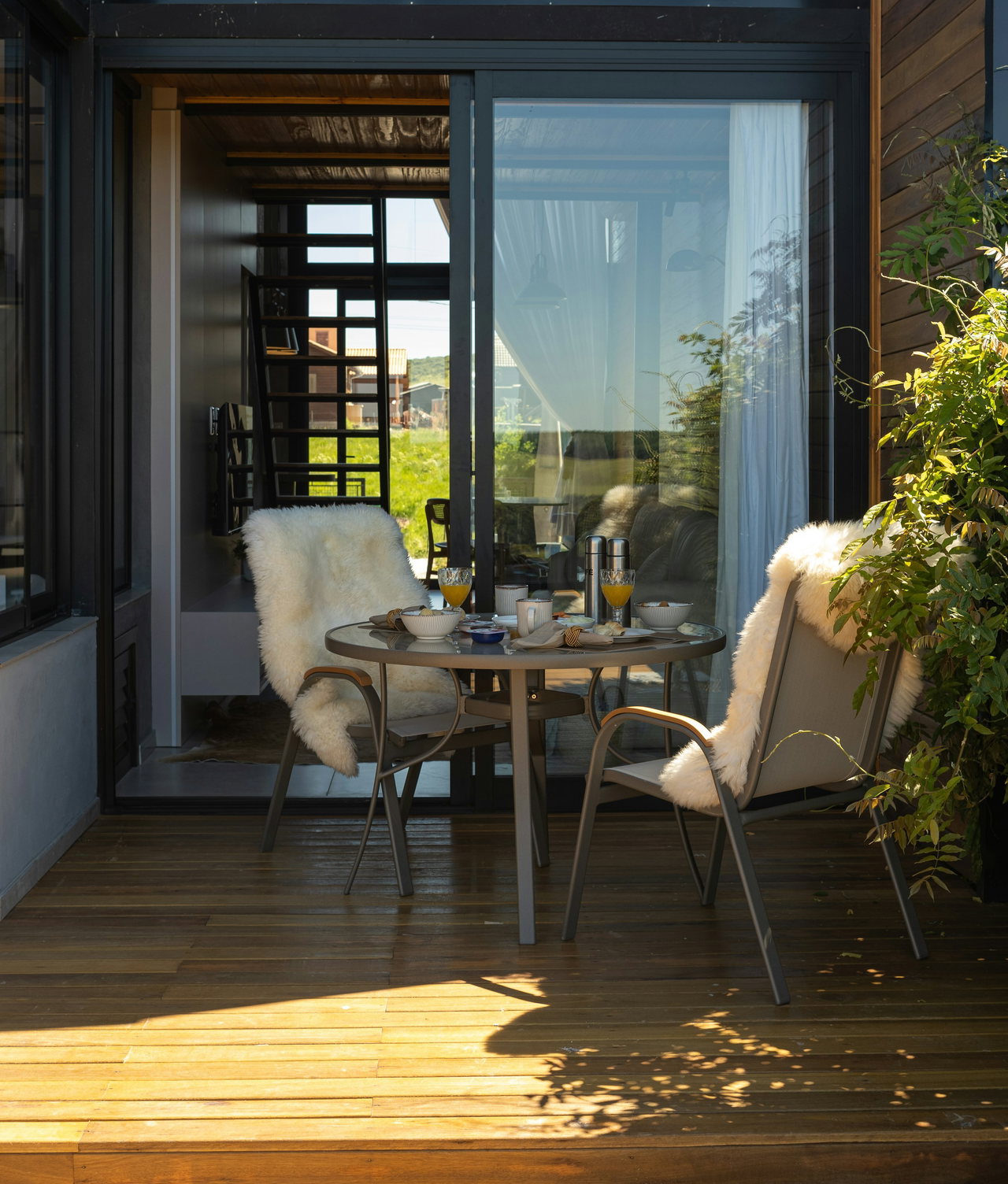 Sonnige Loggia mit Holzdielen, rundem Frühstückstisch und zwei Stühlen mit Lammfellen vor einer offenen Glasschiebetür, rechts Kletterpflanzen.