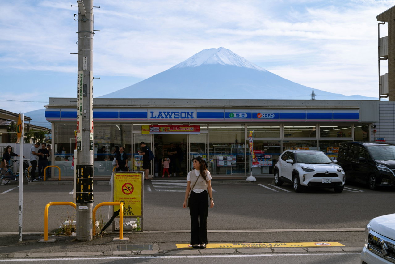 Eine Frau fotografiert an einer Straßenkreuzung den Fuji, der hinter einem Lawson-Laden aufragt