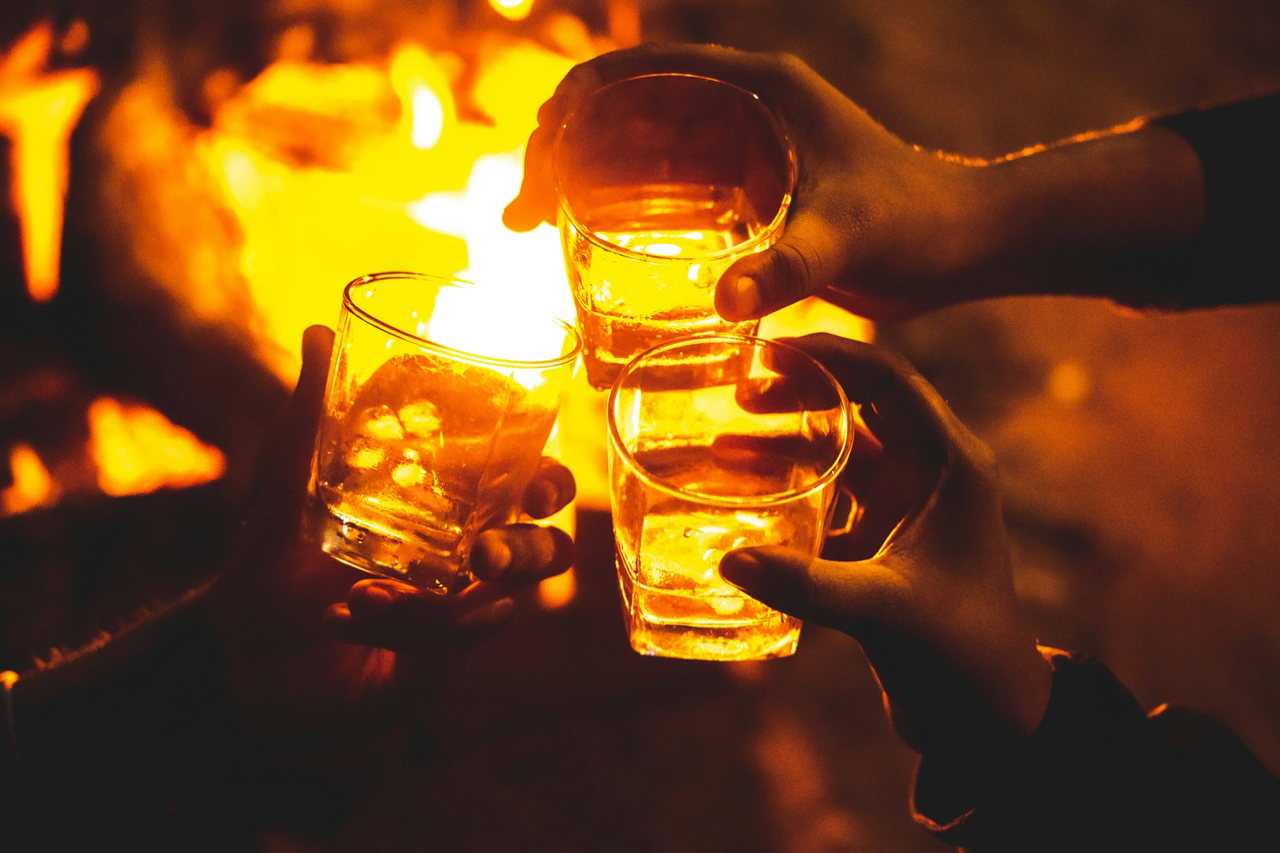 Mehrere Hände halten warm leuchtende Gläser mit heißem Getränk über einem offenen Feuer