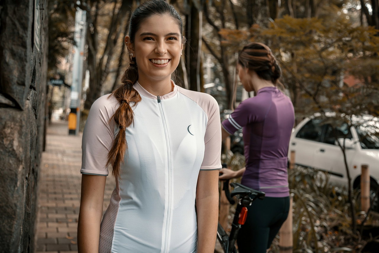 Lachende Frau mit geflochtenem Zopf im hellen Radtrikot auf einem Gehweg, dahinter eine zweite Radfahrerin am Rad