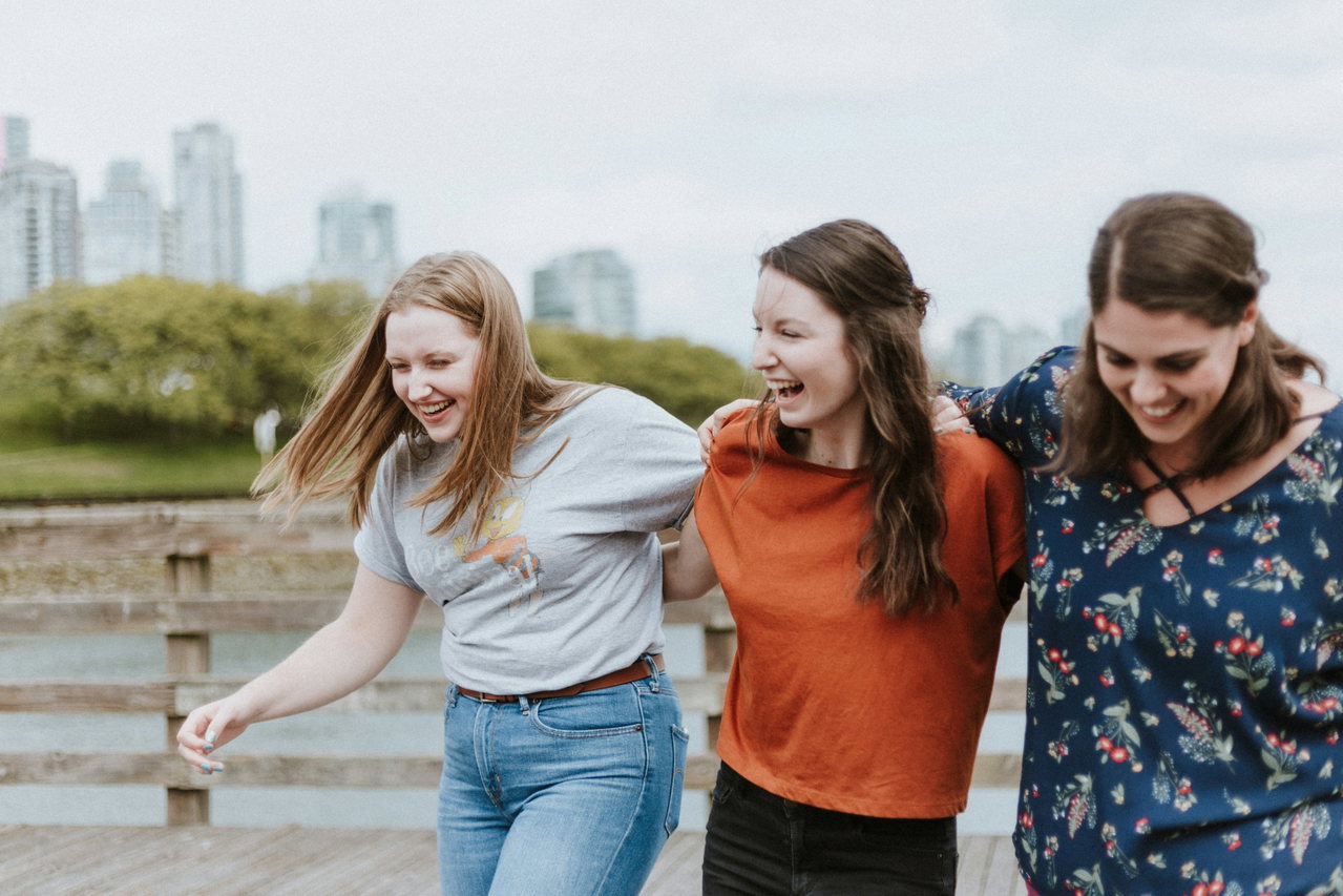 Drei junge Frauen lachen untergehakt im Freien, im Hintergrund unscharf Bäume und Hochhäuser