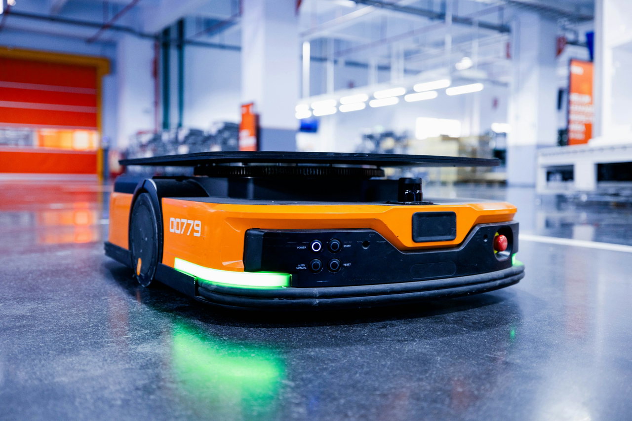 Orangefarbener autonomer mobiler Roboter fährt mit grünem Lichtband durch eine helle Lagerhalle