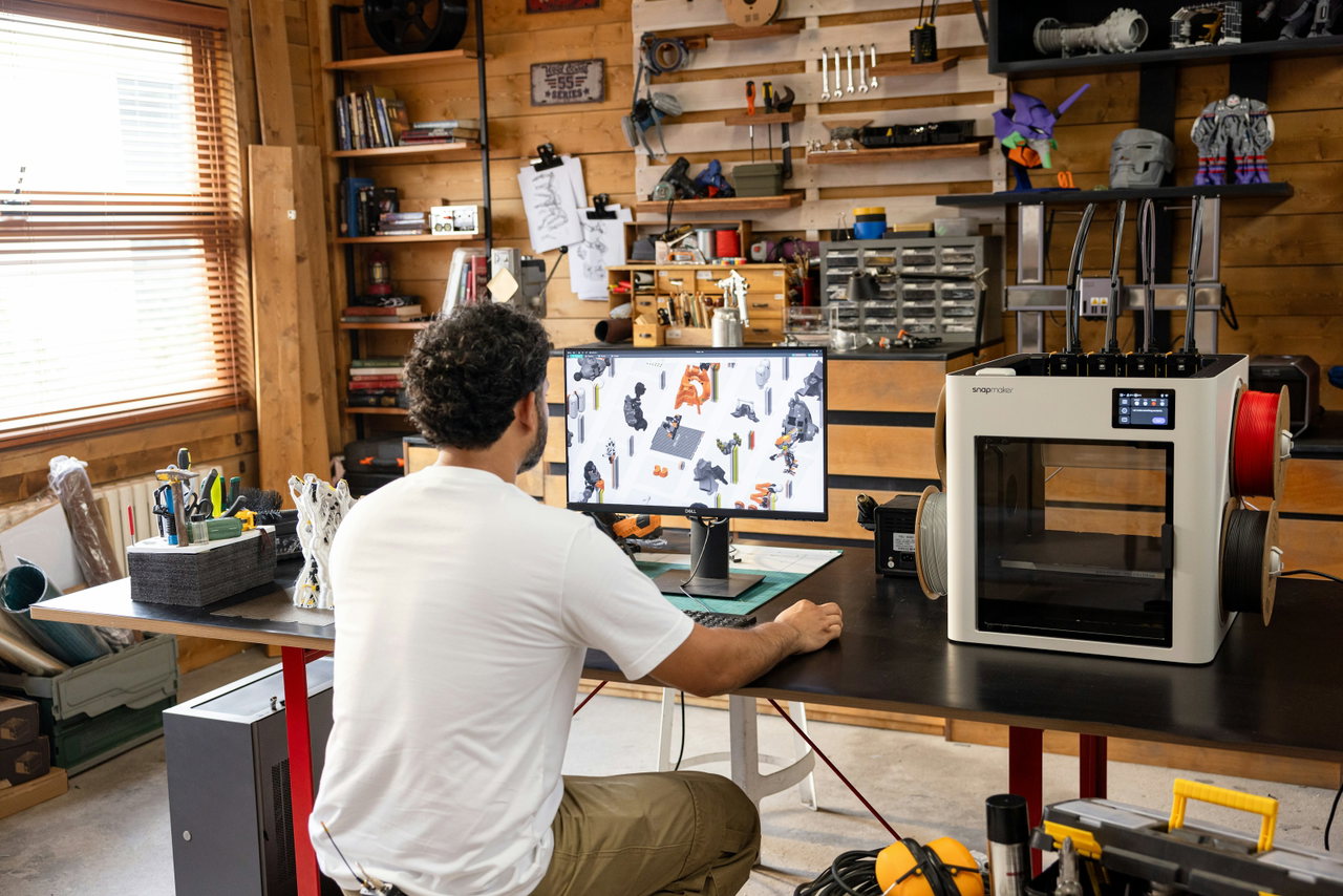Man working at a computer in a workshop using design software, with a 3D printer beside him