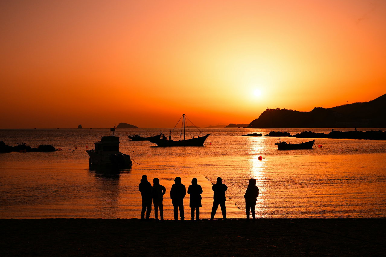 Eine Gruppe steht bei Sonnenuntergang als Silhouette am Meer, davor Boote auf glattem Wasser