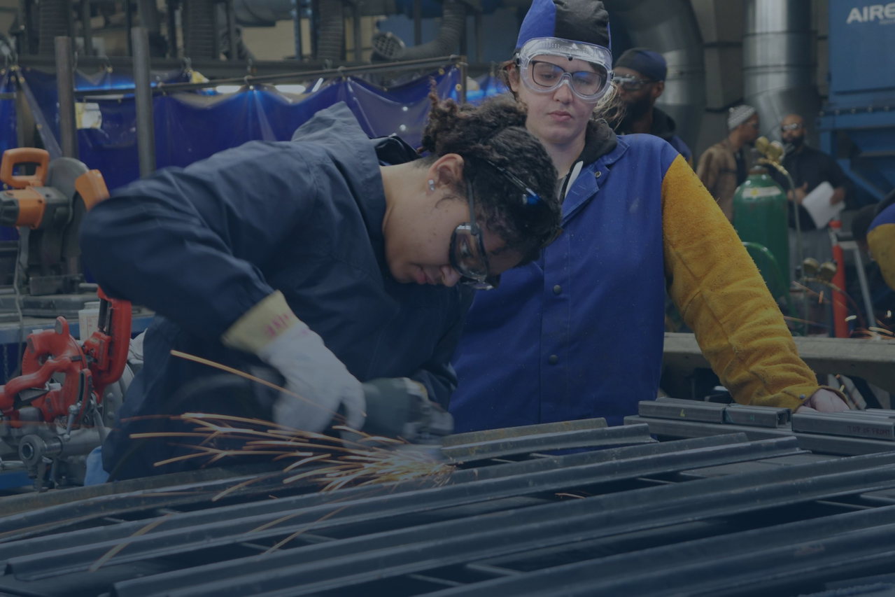 Junge Auszubildende arbeitet mit Schutzbrille und Ausbilderin gemeinsam an einem Metallbauteil, Funken sprühen
