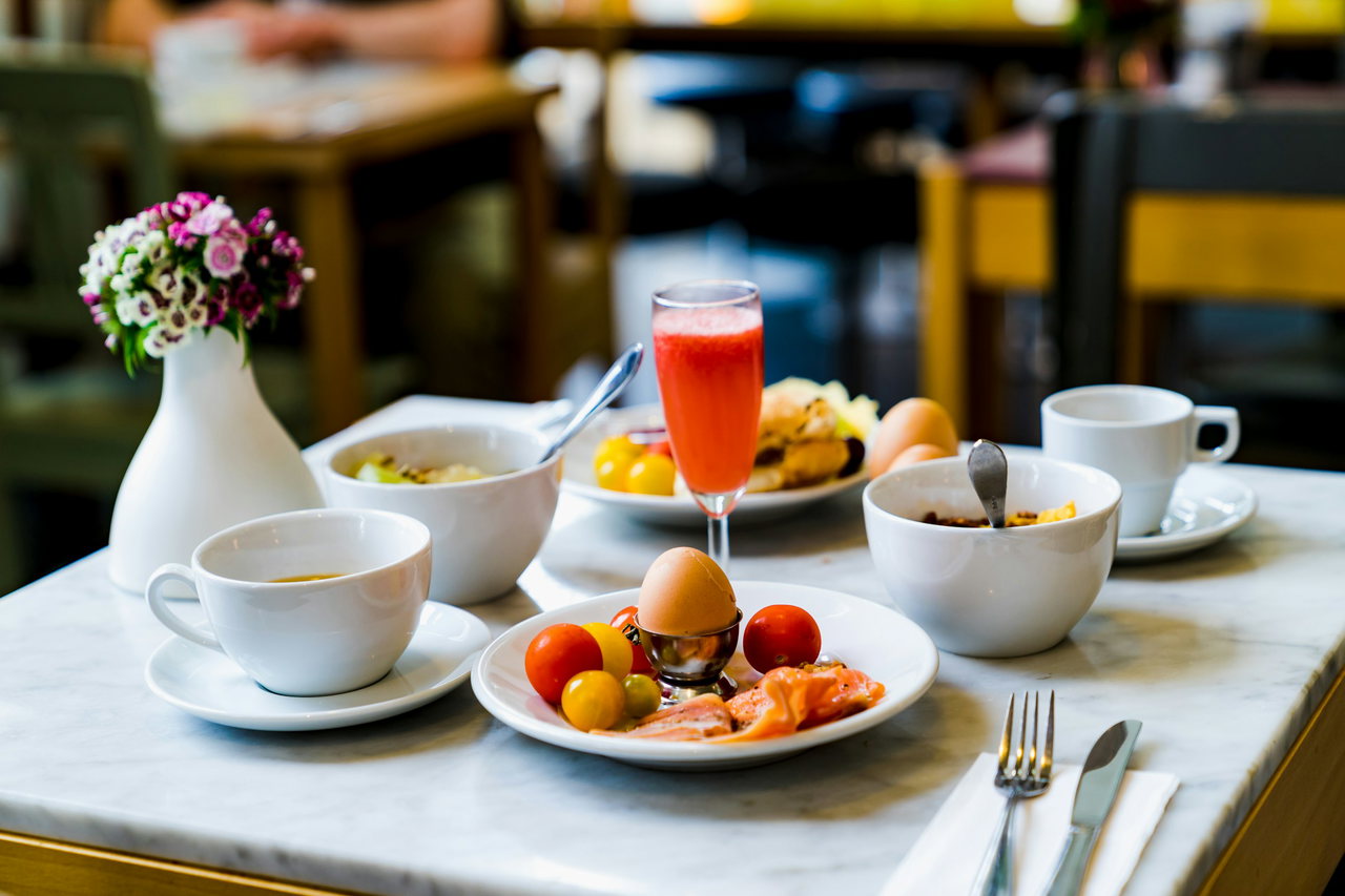 Frühstückstisch mit Marmorplatte: Ei im Eierbecher, Tomaten, Lachs, Müslischalen, Tassen, Saftglas und Blumenvase