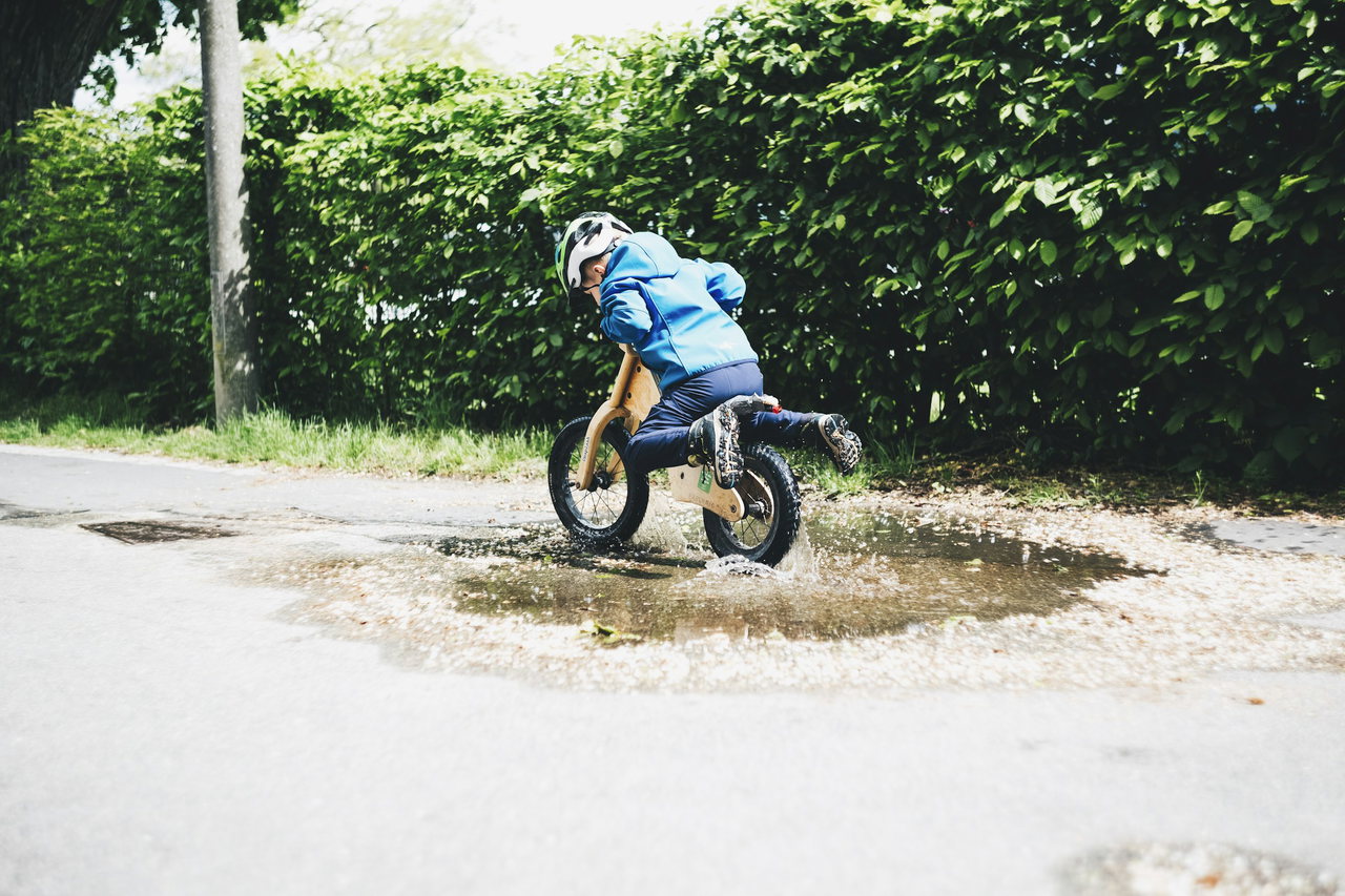 Kind mit Helm und blauer Jacke fährt auf einem Holzlaufrad durch eine spritzende Pfütze