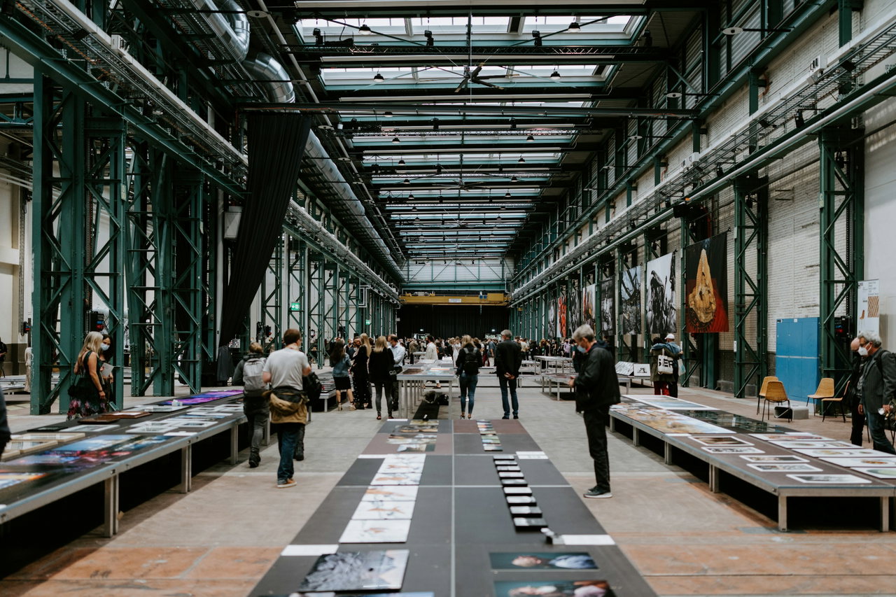 Weite Messehalle mit grünen Stahlträgern, Besuchenden zwischen Ausstellungstischen und Tageslicht von oben