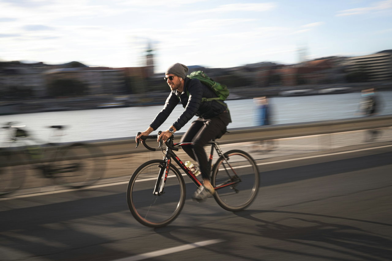 Radfahrer mit Rucksack fährt am Flussufer entlang, die Stadt im Hintergrund ist durch die Bewegung verwischt