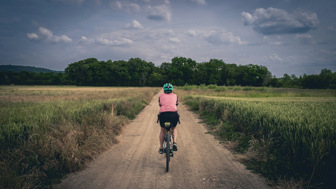 Person mit mintgrünem Helm und rosa Trikot fährt auf einem sandigen Feldweg zwischen Wiesen unter wolkigem Himmel davon