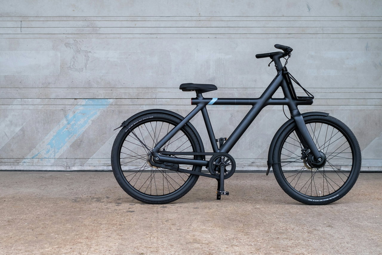 Mattschwarzes Stadt-E-Bike mit Schutzblechen und kleinem Frontgepäckträger steht seitlich vor einer grauen Betonwand