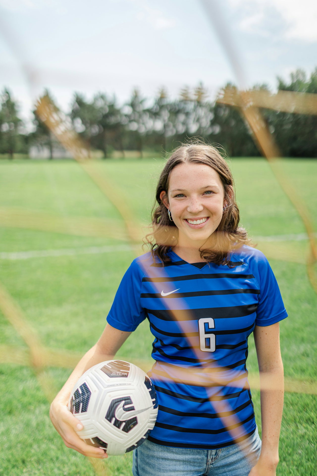 Junge Fußballerin im blau gestreiften Trikot hält lächelnd einen Ball vor einem Tor mit Netz