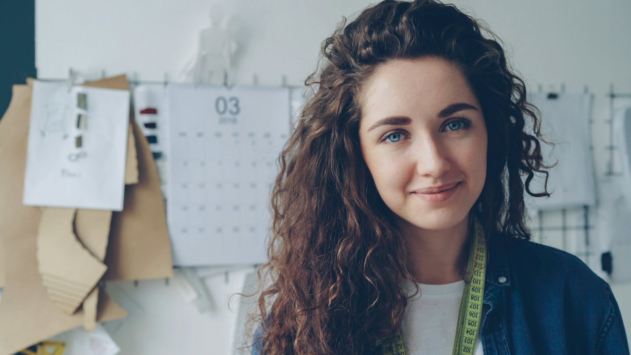 Junge Frau mit langen dunklen Locken, Jeansjacke und Maßband um den Hals blickt freundlich in die Kamera