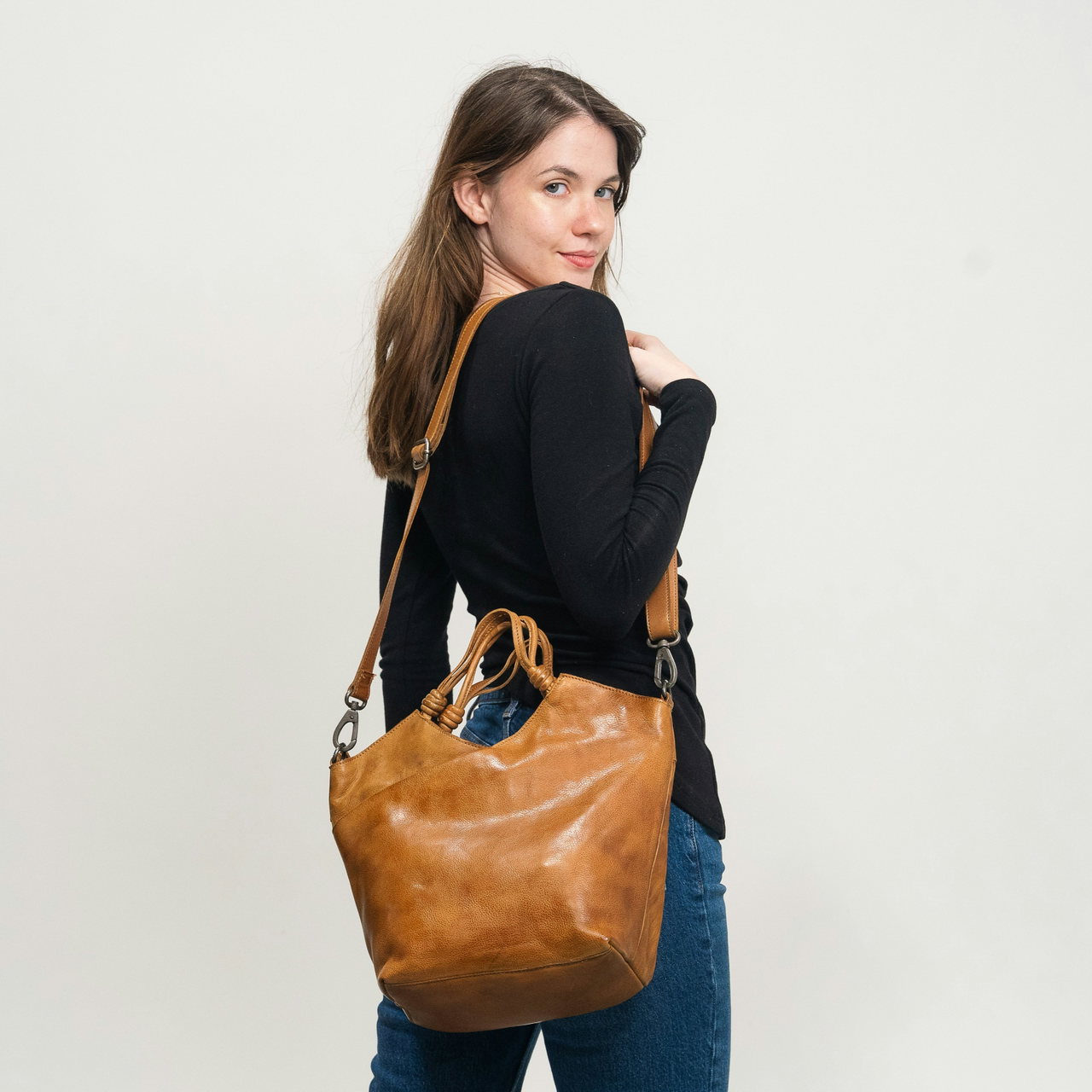 Frau mit Blick über die Schulter trägt eine braune Ledertasche mit Riemen vor hellgrauem Hintergrund