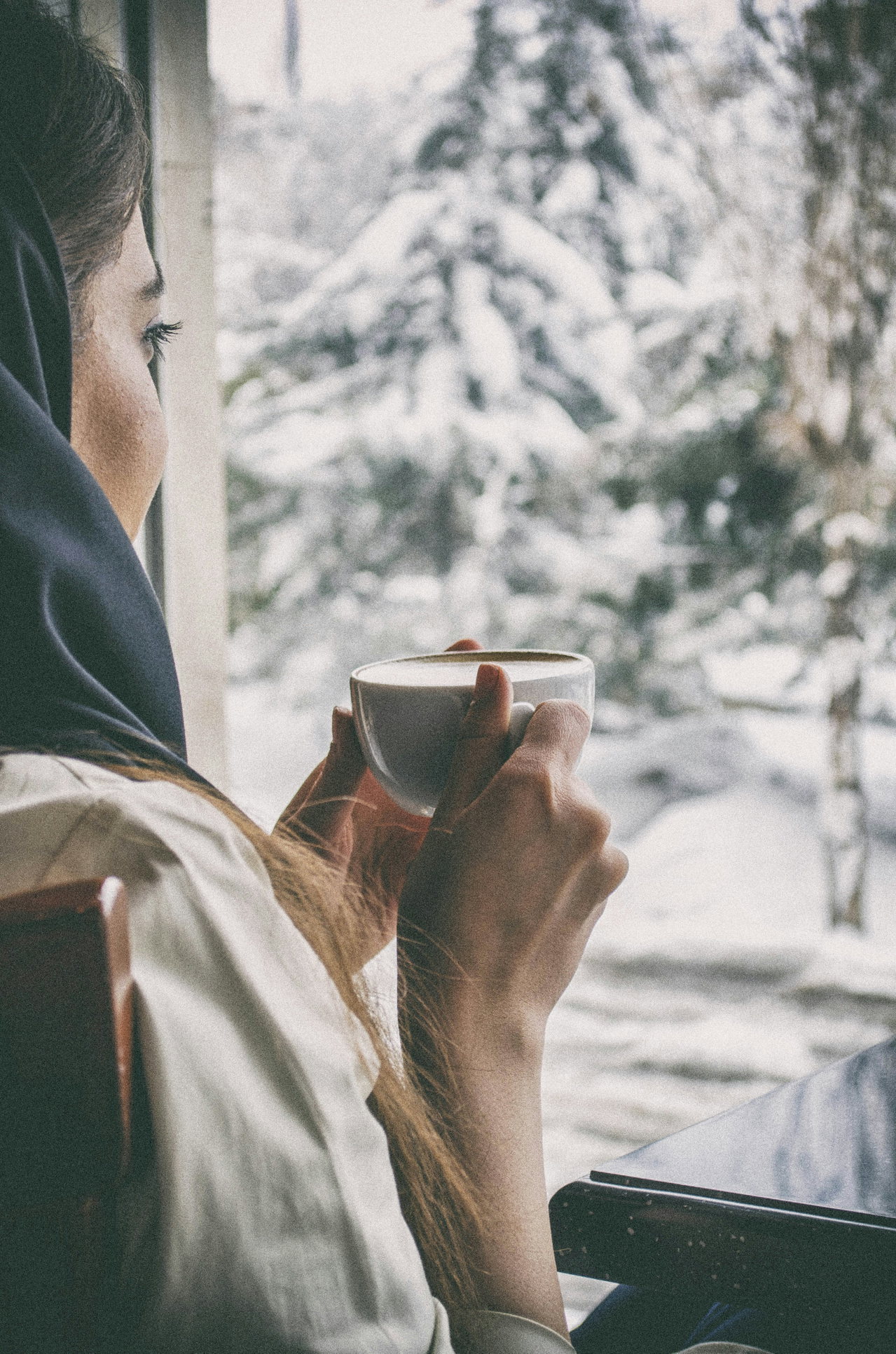 Frau in Schal hält eine Tasse am Fenster, draußen verschneite Fichten