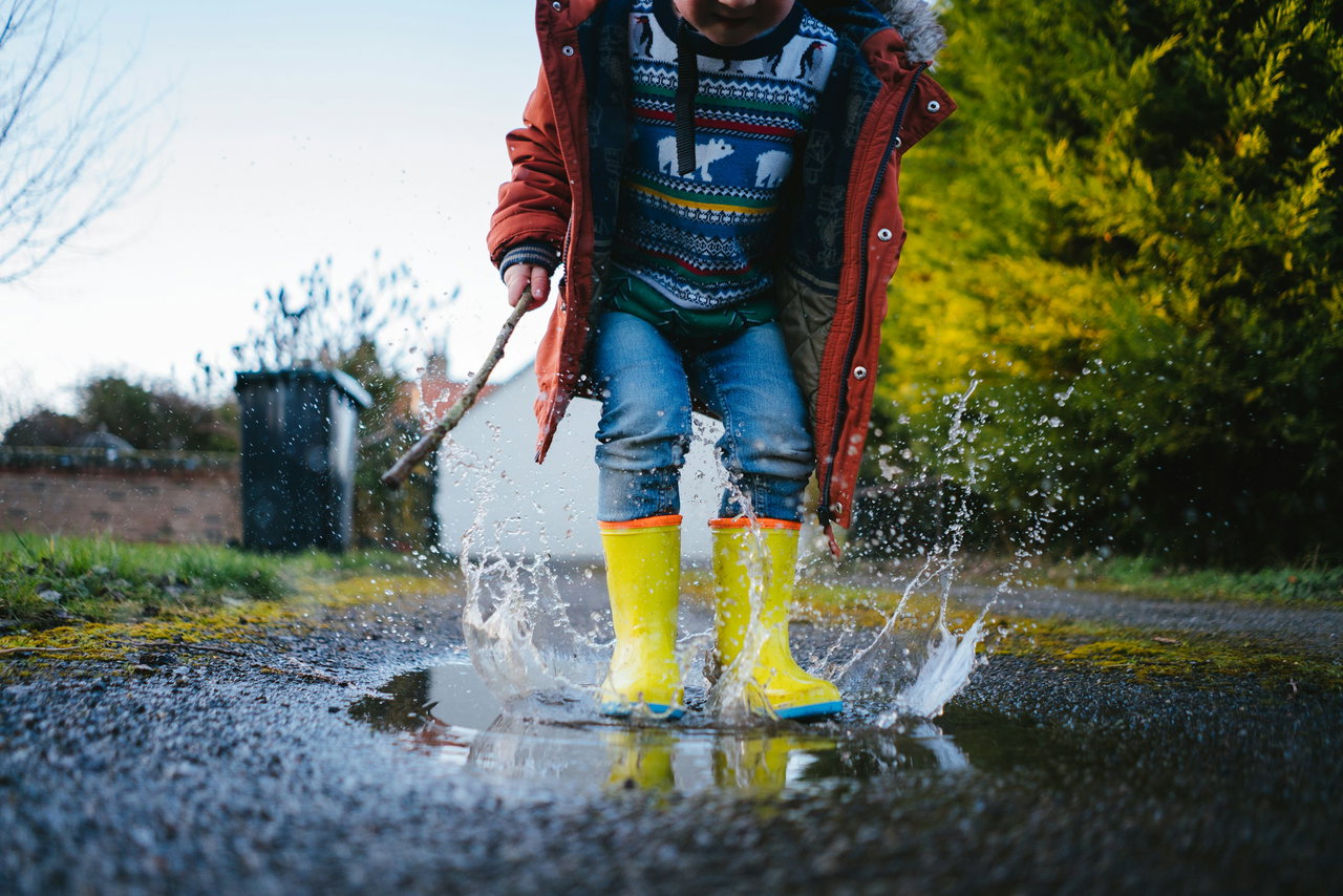 Kind in gelben Gummistiefeln, Jeans und Jacke springt in eine Pfütze, das Wasser spritzt, Gesicht nicht sichtbar