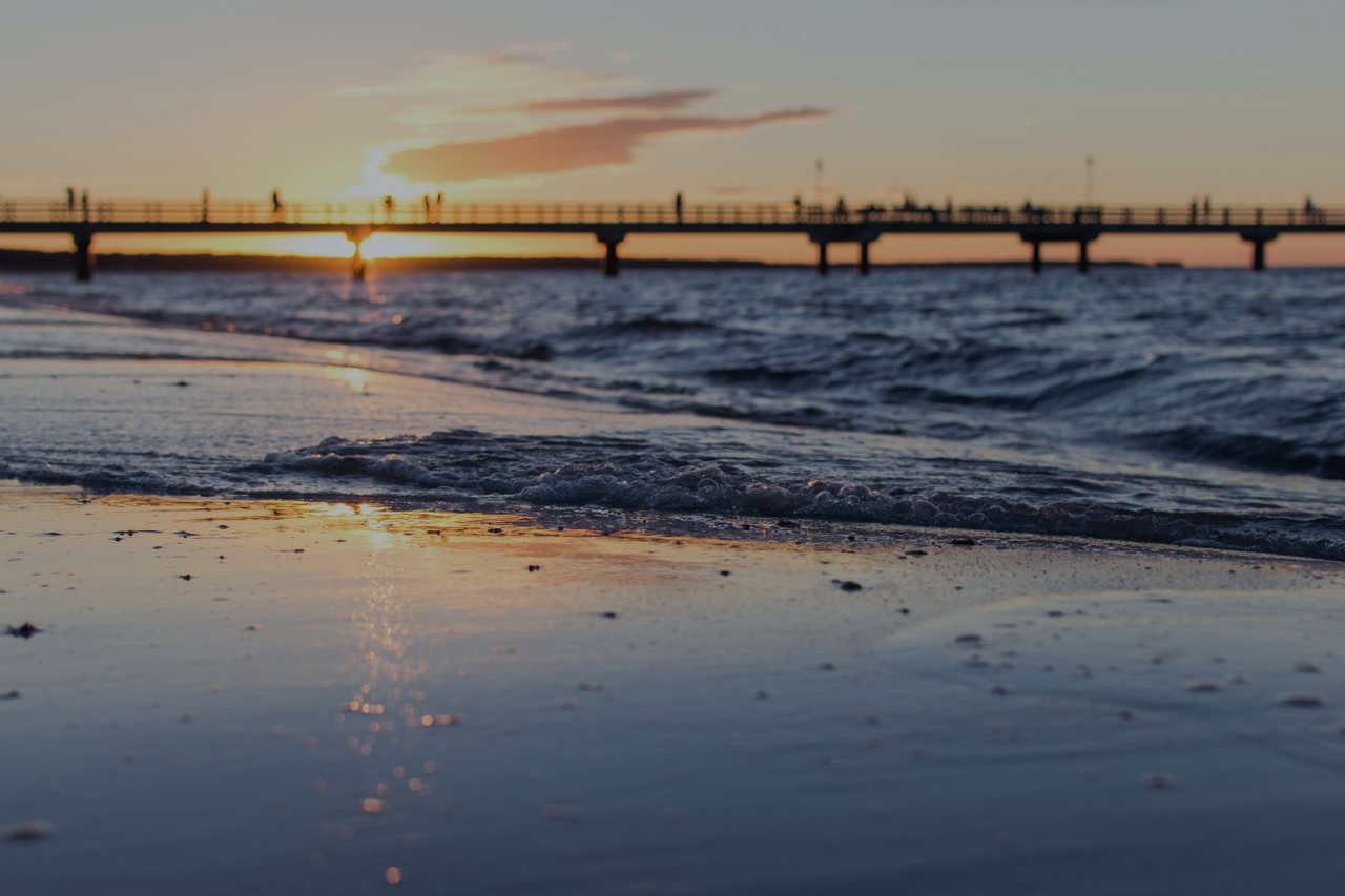 Eine Seebrücke führt im Abendlicht aufs Meer hinaus, die Wellen laufen golden schimmernd auf den Strand