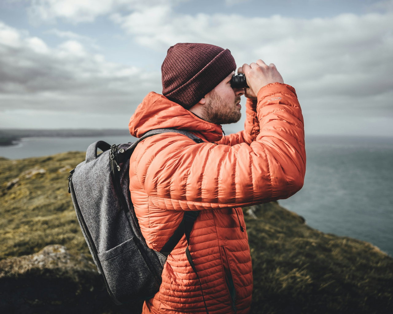 Mann mit weinroter Mütze und orangefarbener Daunenjacke blickt an einer Steilküste durch ein Fernglas in die Ferne