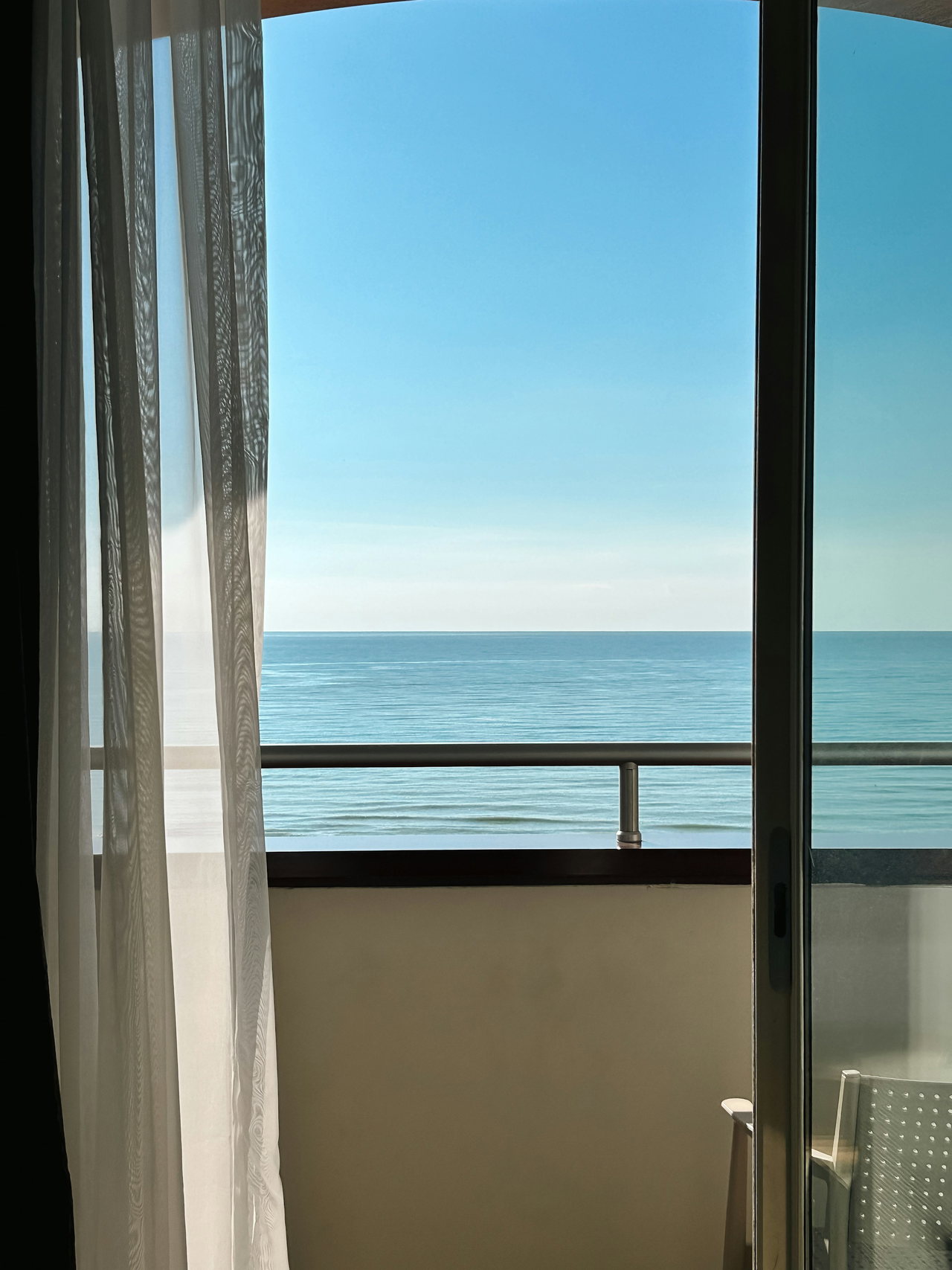 Blick durch die geöffnete Balkontür mit hellem Vorhang auf das ruhige, blaue Meer unter wolkenlosem Himmel
