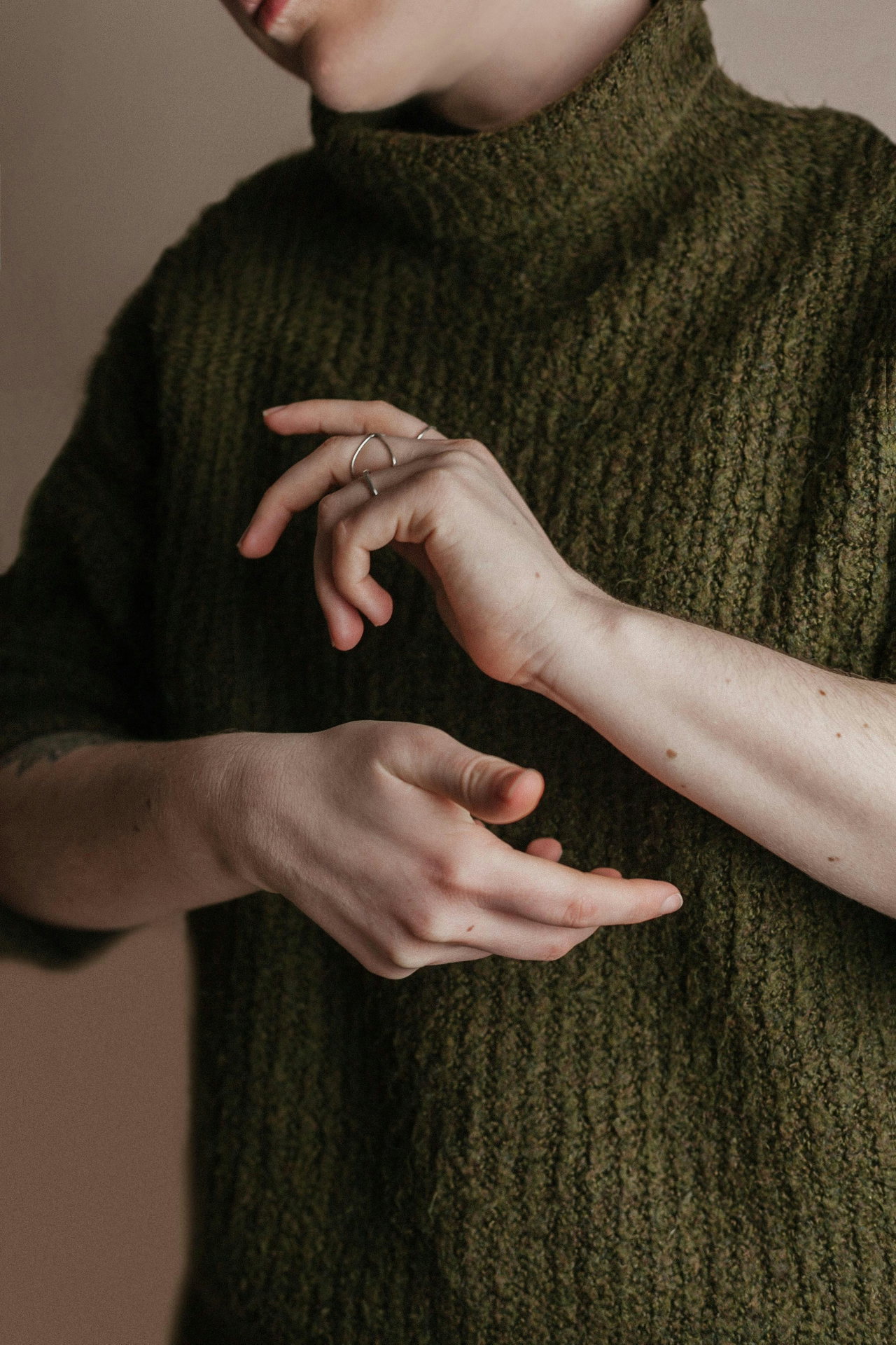 Nahaufnahme: Hände vor einem oliv-grünen Grobstrick-Pullover mit hohem Kragen, ruhiges Studiolicht