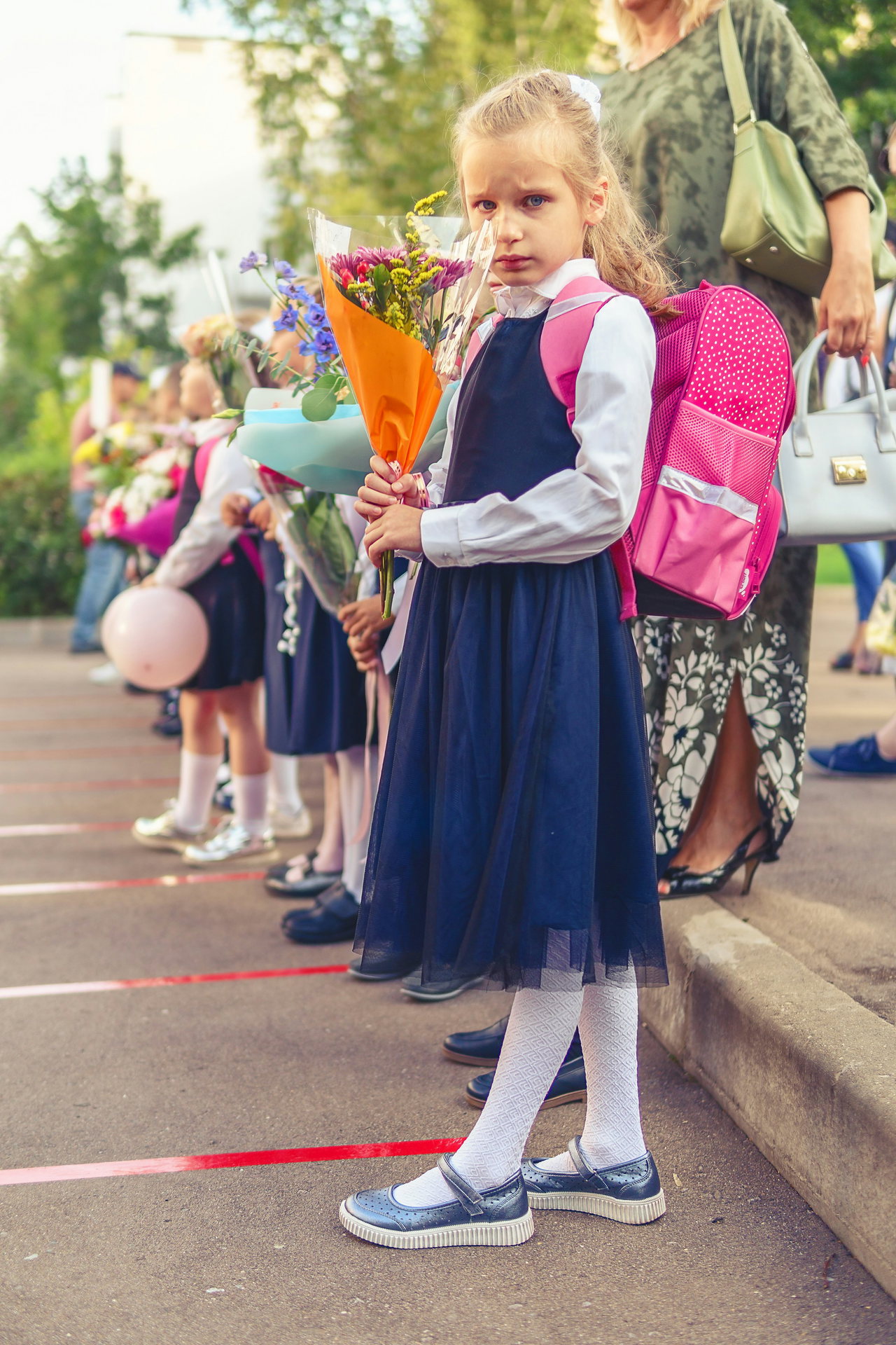 Mädchen in Schulkleidung mit rosa Schulranzen und Blumenstrauß steht am ersten Schultag in einer Reihe mit anderen Kindern