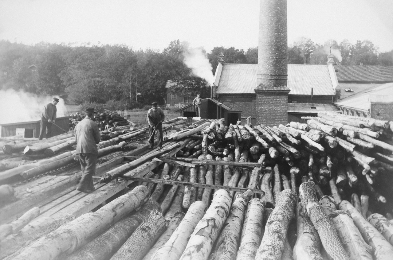 Historisches Schwarz-Weiß-Foto eines Sägewerks: Baumstämme auf dem Holzplatz, dahinter ein Schornstein