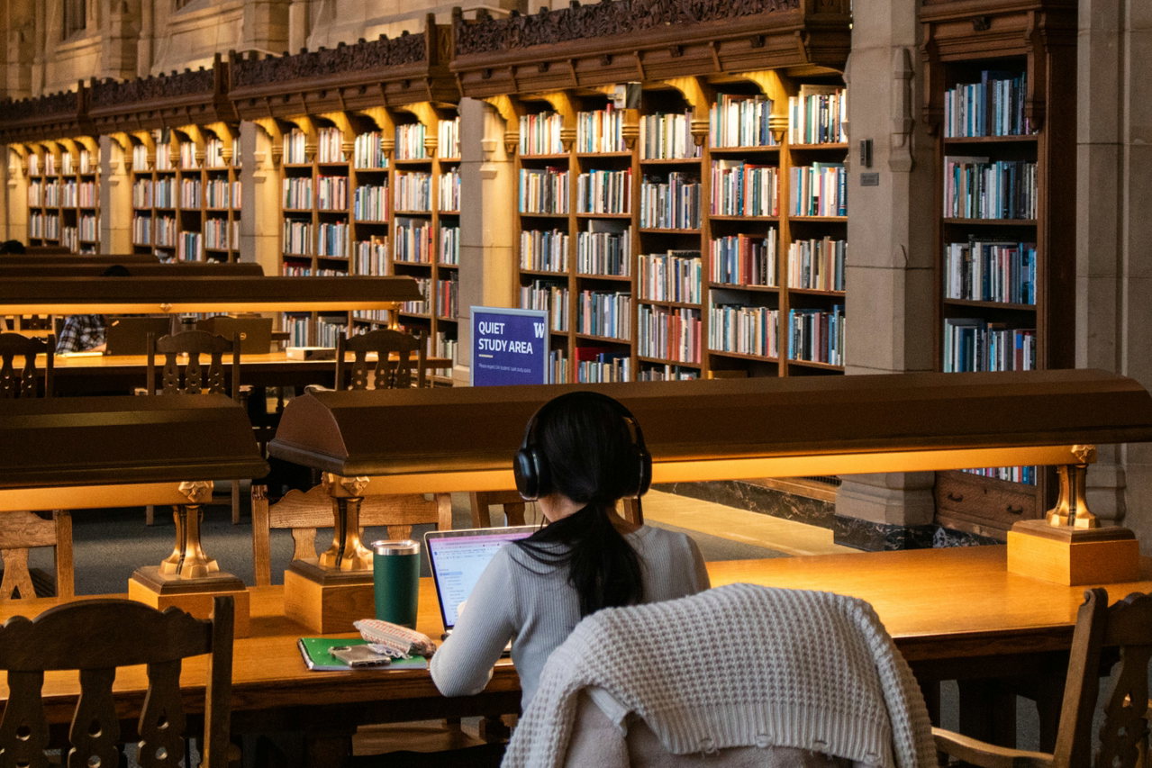 Junge Frau mit Kopfhörern lernt am Laptop an langen Holztischen einer großen, warm beleuchteten Bibliothek